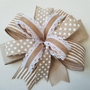 May include: A decorative bow crafted from tan and white ribbon featuring a mix of polka dot and striped patterns. The center of the bow is accented with a delicate white lace trim that edges the loops and tails, creating a layered appearance.