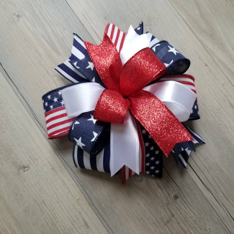 Op de afbeelding: Een decoratieve strik met rode, witte en blauwe linten. De strik heeft een centraal rood glitterlint, witte satijnen linten en linten met Amerikaanse vlagpatronen. Geschikt voor patriottische decoraties.
