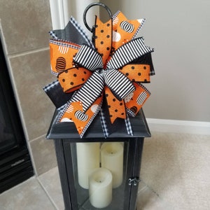 May include: A black lantern with a Halloween-themed bow on top. The bow is made of orange and black ribbon with white polka dots. The lantern has a glass front and is holding three white candles.