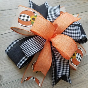 Op de afbeelding: Een grote, veelkleurige strik gemaakt van oranje, zwart en wit lint. De strik heeft een zwart-wit geruit patroon, een effen oranje lint en een beige lint met een pompoenprint.