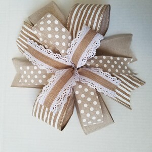 May include: A decorative bow crafted from multiple ribbons featuring a mix of tan burlap, white polka dots, and white vertical stripes. The design includes layers of textured fabric ribbons accented with a delicate white scalloped lace trim.