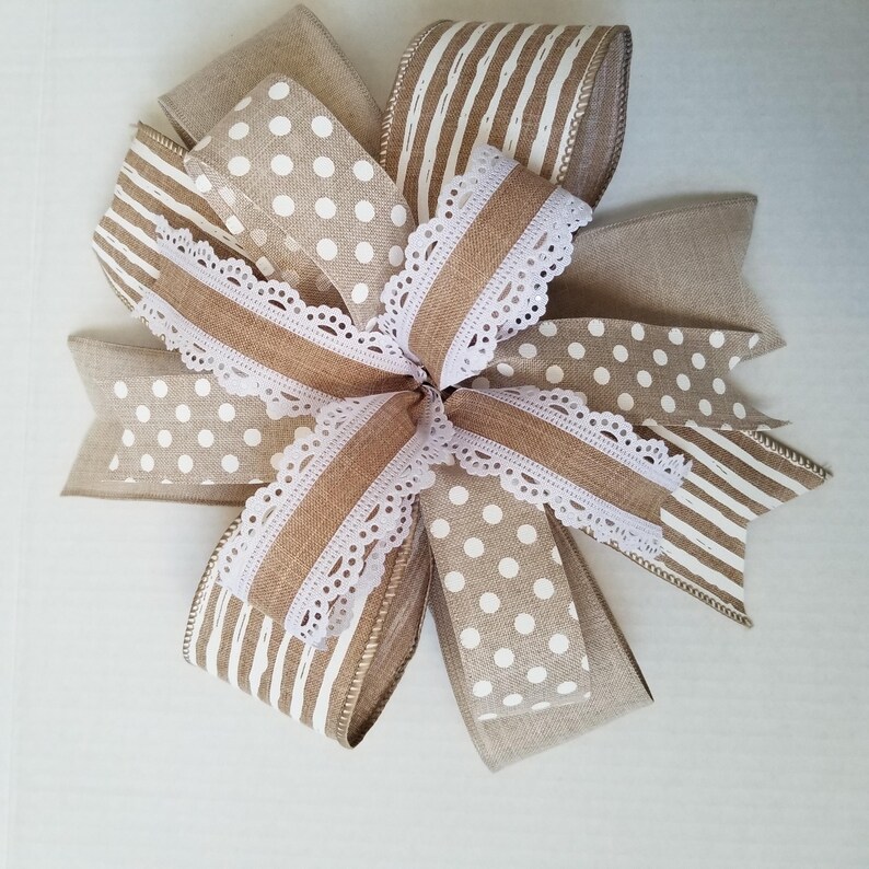 May include: A decorative bow crafted from multiple ribbons featuring a mix of tan burlap, white polka dots, and white vertical stripes. The design includes layers of textured fabric ribbons accented with a delicate white scalloped lace trim.