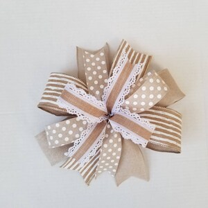 May include: A decorative bow constructed from multiple layers of ribbon in tan and white. The design features a mix of patterns including white polka dots on a tan background, horizontal white stripes on tan, and solid tan, accented with white lace trim.