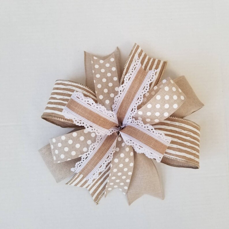 May include: A decorative bow constructed from multiple layers of ribbon in tan and white. The design features a mix of patterns including white polka dots on a tan background, horizontal white stripes on tan, and solid tan, accented with white lace trim.