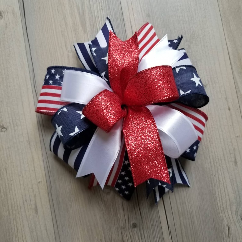 Op de afbeelding: Een decoratieve strik met patriottische kleuren. De strik is gemaakt van verschillende linten, waaronder rode glitter, wit satijn en linten met Amerikaanse vlag- en sterpatronen. Geschikt voor kransen en andere knutselwerkjes.