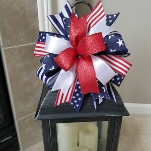 Op de afbeelding: Een decoratieve lantaarn bekroond met een patriottische strik. De strik heeft rood glitterlint, wit satijnlint en linten bedrukt met de Amerikaanse vlag en sterren. De lantaarn is zwart met drie witte kaarsen binnenin.