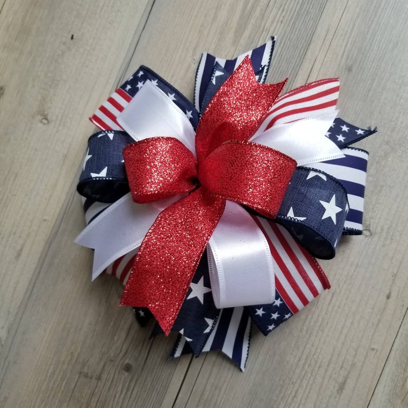 Op de afbeelding: Een decoratieve strik met rode, witte en blauwe linten. De strik heeft een centraal rood glitterlint, witte linten en linten met een sterren- en strepenpatroon. Geschikt voor knutselen, met een patriottisch thema.