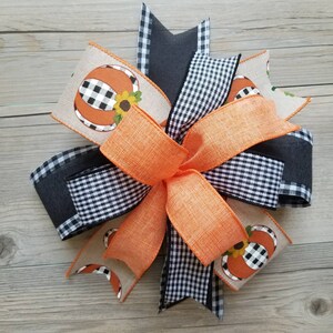 Op de afbeelding: Een grote, meerlagige strik gemaakt van zwart-wit buffalo check lint, oranje jute lint en beige jute lint met een zwart-wit buffalo check pompoenontwerp.
