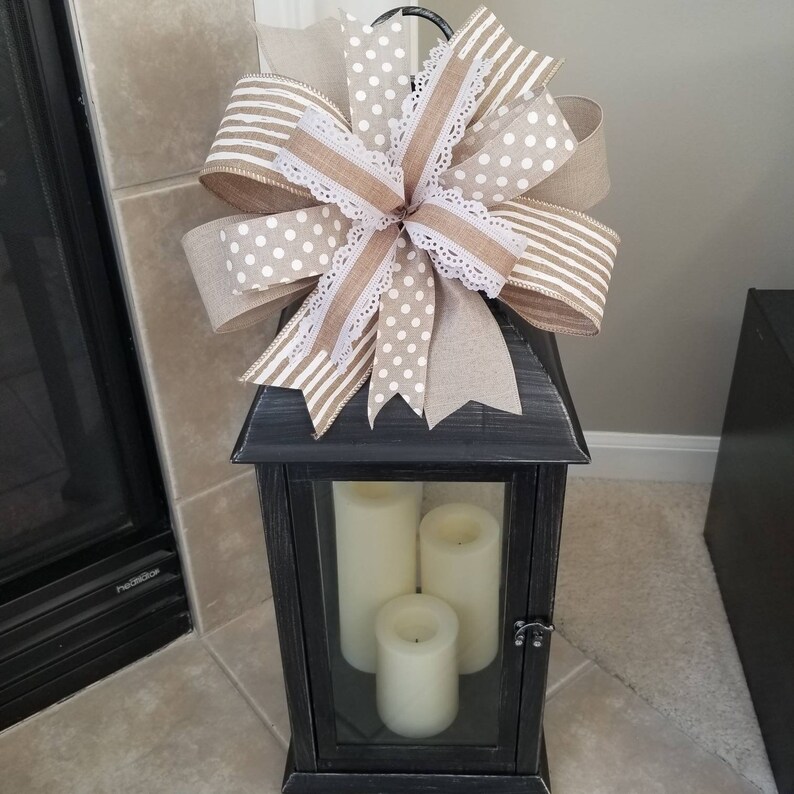 May include: A black lantern with a distressed finish containing three cream-colored pillar candles. A large decorative bow sits on top, crafted from tan burlap, white polka-dot fabric, striped ribbon, and white lace trim with scalloped edges.