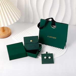 May include: A collection of dark green jewelry packaging, including a gift bag, boxes, and a small pouch, all branded with "PUPPYSEW" in gold. The image also shows a pair of pearl earrings on a green card, indicating a focus on jewelry and gifts.