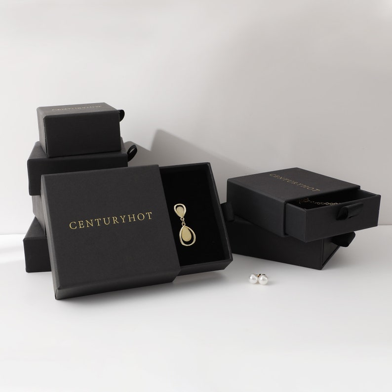 May include: Black jewelry boxes with the text "Centuryhot" printed in gold on the front. The boxes are stacked on top of each other. The top box is open and contains a gold teardrop-shaped earring.