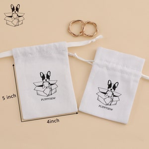 May include: Two white cotton drawstring bags with a black and white illustration of a dog in a box. The larger bag measures 5 inches tall and 4 inches wide. The smaller bag is smaller. Both bags have the text "PUPPYSEW" printed on them.