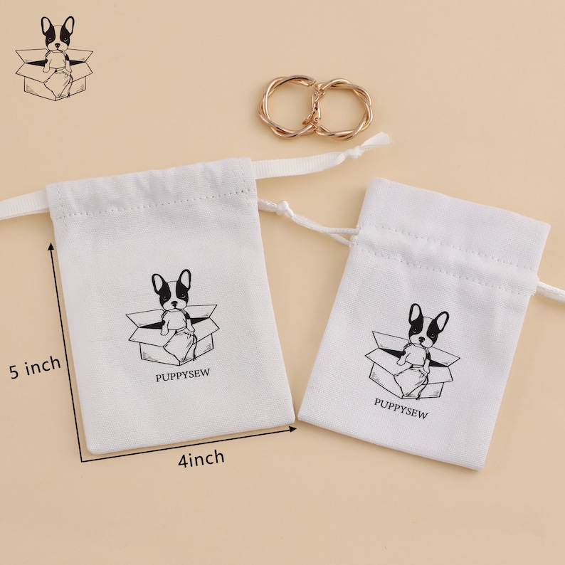 May include: Two white cotton drawstring bags with a black and white illustration of a dog in a box. The larger bag measures 5 inches tall and 4 inches wide. The smaller bag is smaller. Both bags have the text "PUPPYSEW" printed on them.