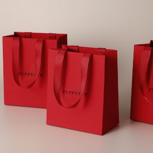 May include: Three rectangular red paper gift bags with matching red ribbon handles. Each bag features the black text PUPPYSEW printed on the front center. The bags are standing upright, showcasing a smooth matte finish and sturdy construction.