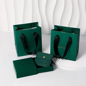 Könnte beinhalten: Smaragdgrüne Geschenktüten und -boxen mit schwarzen Schleifenhenkeln. Die Taschen und Boxen tragen die Aufschrift "PUPPYS" und "PUPPYSEW" in goldfarbenen Buchstaben. Ein kleines grünes Täschchen mit goldenem Knopf ist ebenfalls zu sehen.