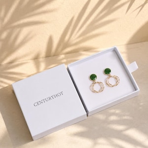 Puede incluir: Un par de pendientes dorados con un perno de piedra redonda de color verde oscuro conectado a un racimo colgante de tres anillos dorados finos y entrelazados, presentados en una caja cuadrada blanca con el texto CENTURYHOT impreso en la tapa.