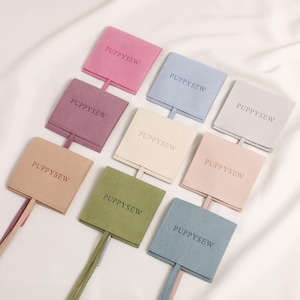 Peut inclure: Collection de neuf pochettes carrées en tissu de couleurs pastel, chacune imprimée du mot "PUPPYSEW". Chaque pochette est dotée d'une fine bande de tissu assortie. Les couleurs incluent le rose, le bleu, le violet, le beige, le vert et la crème.