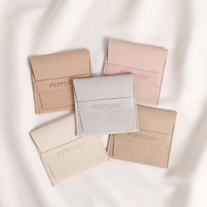 May include: A collection of five small, square pouches in various neutral colors: beige, light pink, and gray. Each pouch has a flap closure and a band across the front with the word "PUPPYSEW" embossed. The pouches are arranged on a white surface.