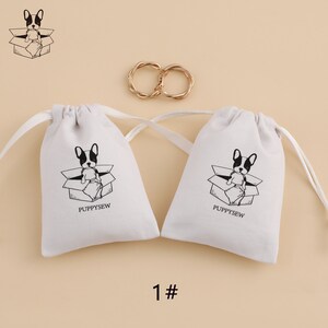 May include: Two white cotton drawstring bags with black and white illustrations of a dog sitting in a box. The text "PUPPYSEW" is printed on each bag.