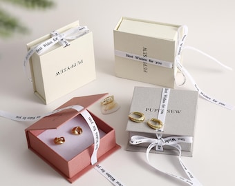 May include: Four gift boxes with white satin ribbon tied in a bow. The boxes are all different colors: white, pink, gray, and a light beige. The boxes are all square and have the text "Best Wishes for you" printed on the ribbon. The boxes are all labeled with the text "PUPPYSEW".