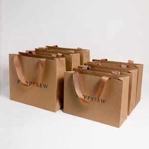 May include: A collection of rectangular brown paper gift bags featuring the text POPPYSEW printed in black on the front. Each bag is equipped with two brown satin ribbon handles for carrying, arranged in a staggered formation on a plain surface.