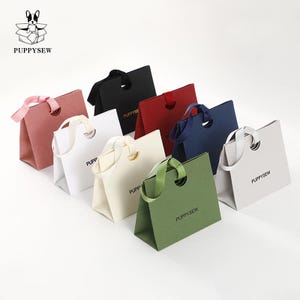 May include: A selection of colourful gift bags in shades of pink, white, black, red, navy, green, and grey. Each bag has a unique handle design and the brand name "PUPPYSEW" printed on the side. The bags are arranged on a white surface.