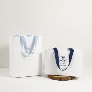 May include: Two white paper gift bags with blue and navy blue ribbon handles. The bags have a logo with a cartoon dog in a box and the text "Puppysew".