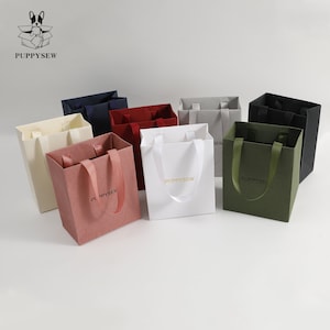 May include: A collection of colorful gift bags in various shades, including pink, white, and navy. Each bag features a matching ribbon handle and the brand name "PUPPYSEW" printed on the front. The bags are arranged on a white surface.
