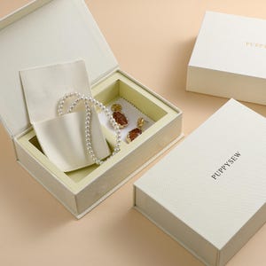 May include: Ivory jewelry box containing a pearl necklace and gold-coloured earrings with brown stones. A small ivory pouch holds the necklace. The box is on a light beige surface.