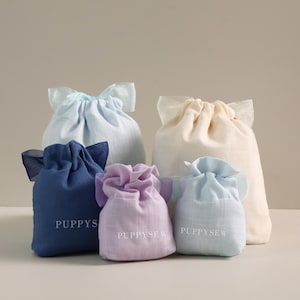 May include: An assortment of drawstring pouches in various pastel colors, including light blue, lavender, and cream. The pouches have a soft, textured appearance and are gathered at the top with a ribbon. The word "PUPPYS" or "PUPPYSEW" is printed on some of the pouches.