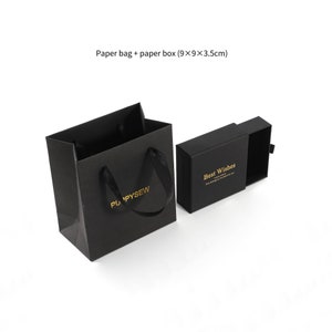 May include: Black paper gift bag with black ribbon handles and gold lettering that says "PU PYSEW". A black paper box with a drawer is open and says "Best Wishes" in gold lettering. The bag and box are on a white background.