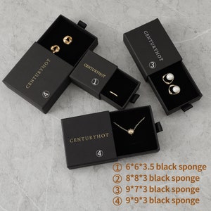 May include: Four black boxes with gold lettering that says "CENTURYHOT" and a number from 1 to 4. Each box contains a different piece of jewelry: a pair of gold earrings, a gold ring, a pair of pearl earrings, and a gold necklace with a star pendant. The boxes are on a gray textured surface. The text below the boxes reads: "① 6*6*3.5 black sponge ② 8*8*3 black sponge ③ 9*7*3 black sponge ④ 9*9*3 black sponge"