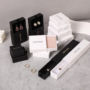 Puede incluir: Una variedad de joyeros en blanco y negro, algunos abiertos para revelar pendientes y collares. Las cajas están etiquetadas con "PUPPYSEW" en letras doradas. También se ven pendientes de aro dorados.