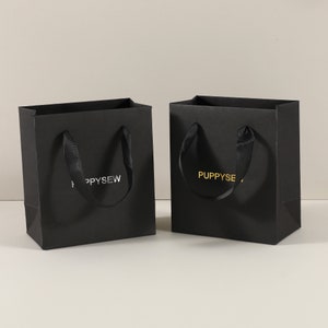 May include: Two black paper gift bags with black ribbon handles. One bag has the text "PUPPYSEW" in silver foil lettering. The other bag has the text "PUPPYSEW" in gold foil lettering.