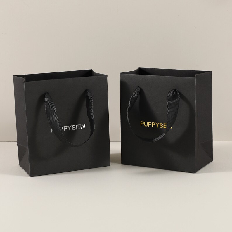 May include: Two black paper gift bags with black ribbon handles. One bag has the text "PUPPYSEW" in silver foil lettering. The other bag has the text "PUPPYSEW" in gold foil lettering.