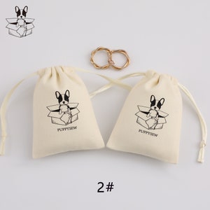 May include: Two small, off-white drawstring bags with black and white illustrations of a dog in a box. The bags have the text "Puppysew" printed on them. The bags are on a white background with a gold ring in the center.