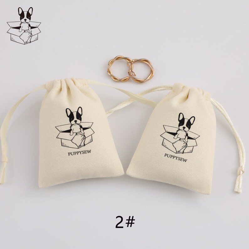 May include: Two small, off-white drawstring bags with black and white illustrations of a dog in a box. The bags have the text "Puppysew" printed on them. The bags are on a white background with a gold ring in the center.