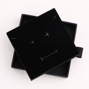 May include: Black velvet jewelry box with a square lid. The lid has a white design of two plus signs and a horizontal line.