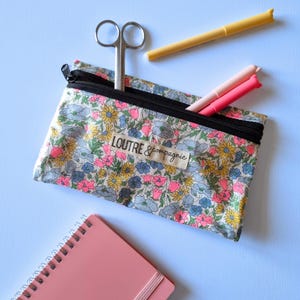 May include: A floral print pencil case with a black zipper, containing scissors and pens. The case has a label that reads "loutre & compagnie". A pink notebook and yellow pen are also visible.