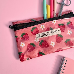 May include: A pink pencil case with a black zipper, filled with pens and scissors. The case is decorated with a strawberry and flower pattern, and the text "LOUTRE & compagnie" is visible.