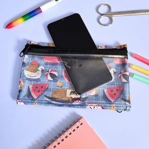 May include: A blue pencil case with a black zipper, featuring a summer-themed pattern of ducks, watermelons, and beach balls. A smartphone is partially visible inside the case, along with a clear pocket. Other stationery items are scattered around.