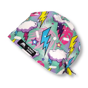 Op de afbeelding: Een groene, paarse, roze en gele gepatroneerde chirurgische scrub-muts met een wit wolken- en bliksemontwerp. De muts heeft een strik aan de achterkant en een label met de tekst "la loutre piqueuse".
