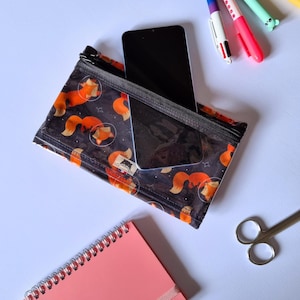 May include: A clear zippered pouch with a black zipper, holding a black smartphone. The pouch is patterned with orange foxes in space suits on a dark blue background. Other items include pens, scissors, and a pink notebook.