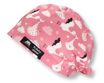May include: Pink surgical scrub hat with a white stork and baby design. The hat has a tie at the back and a label that says "la loutre piqueuse".