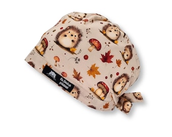 May include: Beige fabric head covering with a repeating pattern of cartoon hedgehogs, red-capped mushrooms, and autumn leaves. The head covering has a tie closure and a small black label with white text.
