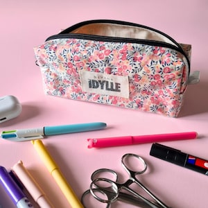 May include: A floral print pencil case with a black zipper and the text "DIVINE IDYLLE" on a rectangular patch. Assorted pens, scissors, and other stationery items are scattered on a pink surface.