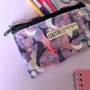 May include: A rectangular pencil case with a black zipper, featuring a purple, pink, and white galaxy design with whales, moons, and stars. The text "LOUTRE & compagnie" is printed on a small label.