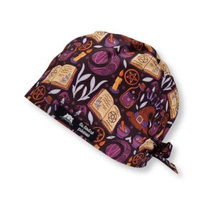 Può includere: Un cappello chirurgico viola scuro con un motivo ripetuto di illustrazioni a tema strega. Il design include libri di incantesimi aperti, candele, bottiglie di pozioni, cappelli da strega e lune crescenti. Il cappello ha una chiusura con lacci.