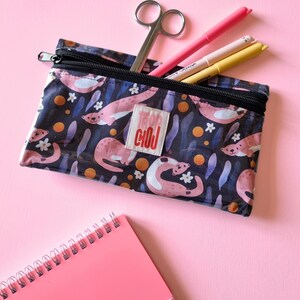 May include: A clear pencil case with a black zipper, filled with pens and scissors. The case features a pattern of pink otters, white flowers, and orange circles on a dark blue background. A pink notepad is in the foreground.