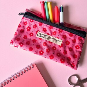May include: A pink floral pencil case with a black zipper, holding colorful pens. The case has a label that reads "LOUTRE & compagnie". A pink notebook and scissors are also visible.